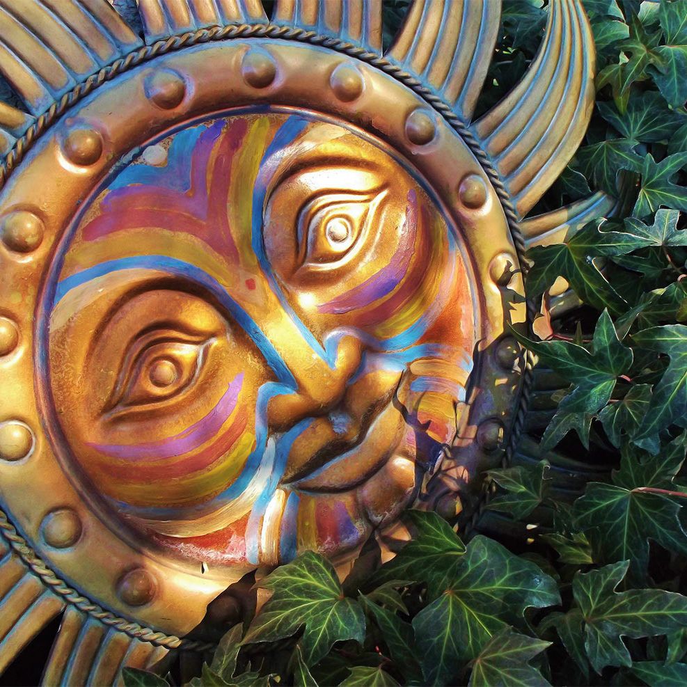 Sun Spirit in the Vines by Usha Lamat, a photograph of a golden painted sun face partially hidden among deep green ivy leaves with radiant symbolic detail